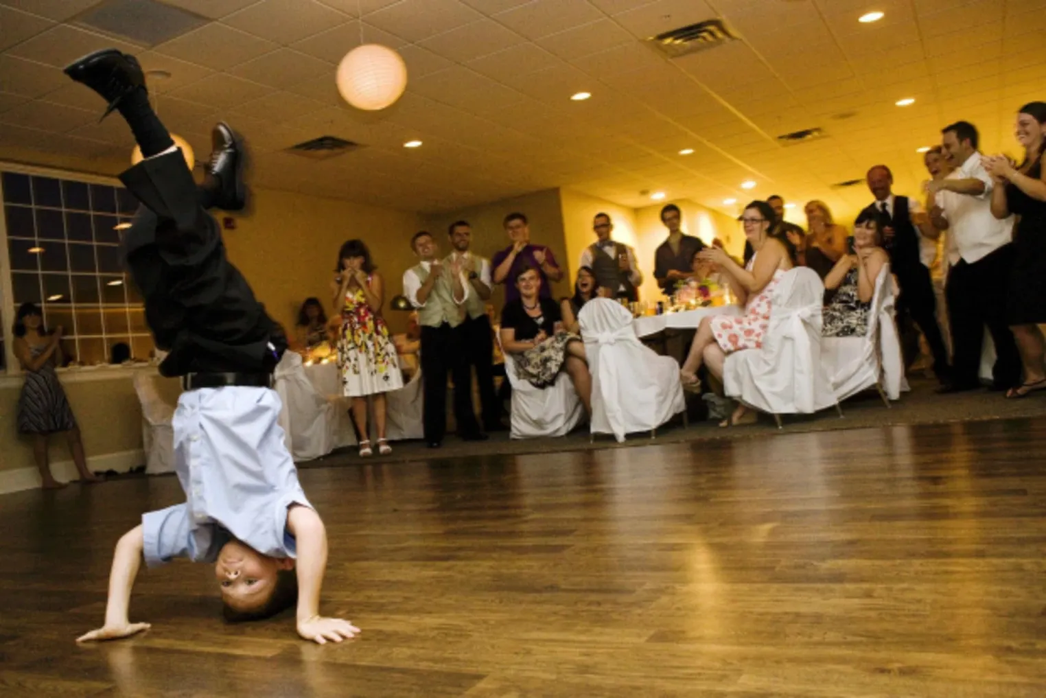 A child on his head break dancing on the dance floor of a wedding reception with a crowd of people cheering in the background